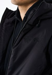 Sort ærmeløs jakke med hætte, lavet af glat stof, med lynlåslukning og broderet logo på brystet, lagdelt over en sort hoodie.