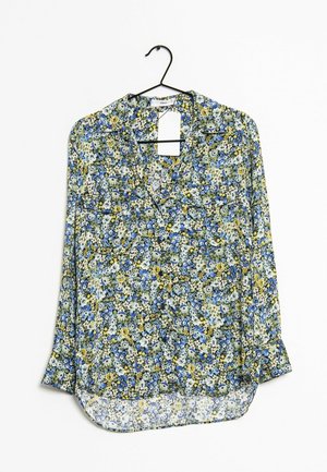 Bloes met lange mouwen en bloemenpatroon in blauw, geel en wit, hangend aan een zwarte hanger tegen een witte achtergrond.