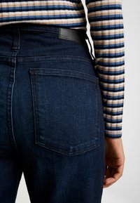 Mörkblå denimjeans har hög midja, bakfickor och en svart läderetikett. En randig långärmad topp är delvis synlig.