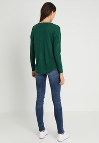 Grünes langärmliges Oberteil mit lockerem Schnitt und rundem Ausschnitt, kombiniert mit eng anliegenden blauen Jeans und weißen Turnschuhen. Einheitlich glatte Textur.