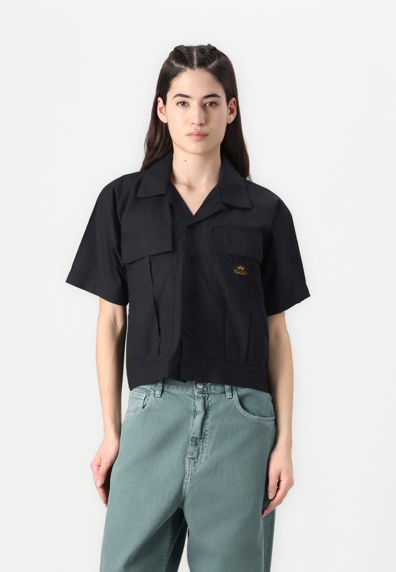 Camisa preta cropped com botão e mangas curtas, apresentando dois bolsos frontais e um detalhe sutil do logo. Combinada com jeans soltos de cor verde-claro.