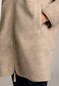 Bež pulover s puhasto teksturo, z zadrgo spredaj, stranskimi žepi in kontrastnim toniranim robom ob robovih.