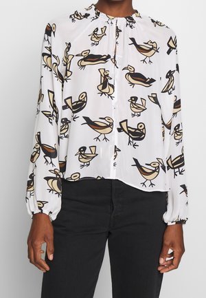 Blusa blanca de manga larga con cuello ruche y estampados abstractos de pájaros en tonos marrón, beige y negro, combinada con pantalones negros.