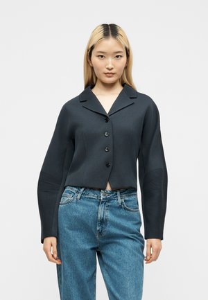 Jeune femme aux cheveux blonds raides porte une veste noire boutonnée et un jean bleu ample à taille haute, debout devant un fond uni.