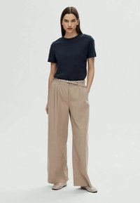Mulher em pé com as mãos nos bolsos, vestindo uma blusa preta de mangas curtas, calças largas bege com cinto e sapatos bege em fundo liso.