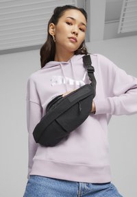 Černá látková crossbody taška se zipovou kapsou, nošená přes světlofialovou mikinu s bílým grafickým designem. Pohodlné džíny.