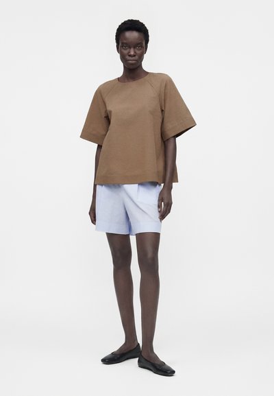 Γυναίκα που στέκεται, φοράει ένα χαλαρό καφέ τοπ, ανοιχτό μπλε σορτς και μαύρα φλατ παπούτσια, μπροστά σε απλό λευκό φόντο.