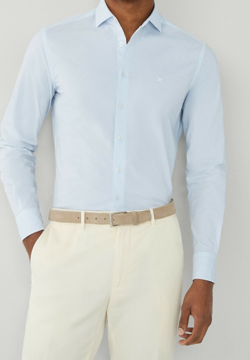 Vaaleansininen pitkähihaisessa nappipaidassa on slim fit -malli, teksturoitu kangas ja pieni brodeerattu logo rinnassa. Beigepohjainen vyö ja kermaiset housut.