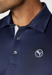 Sötétkék polo ing rövid gallérral, két gombbal és fehér Puma logóval a mellrészen. Az anyag simának és lélegzőnek tűnik.