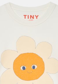 Valkoinen puuvillapaita, jossa on suuri hymyilevä kukkakuva, oranssi keskus ja siniset silmät, merkitty brändillä "TINY" punaisella.