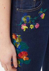 Los jeans de mezclilla azul oscuro presentan un colorido bordado floral en amarillo, rosa, verde y rojo, acentuando los bolsillos y los lados.