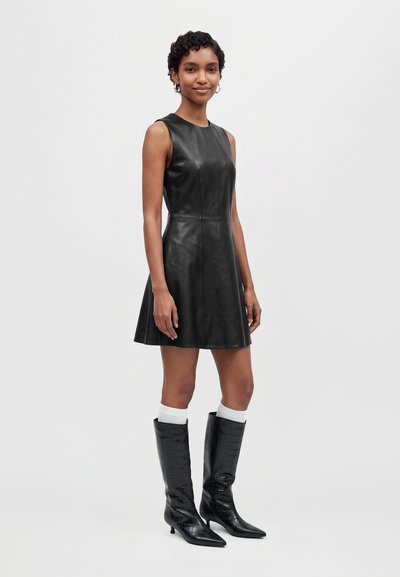 Une femme porte une mini-robe noire en cuir sans manches et de hautes bottes noires sur un fond clair uni.