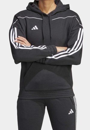 Osoba ubrana w czarną bluzę z kapturem i spodnie Adidas z białymi paskami, z założonymi ramionami, na jasnym, jednolitym tle.