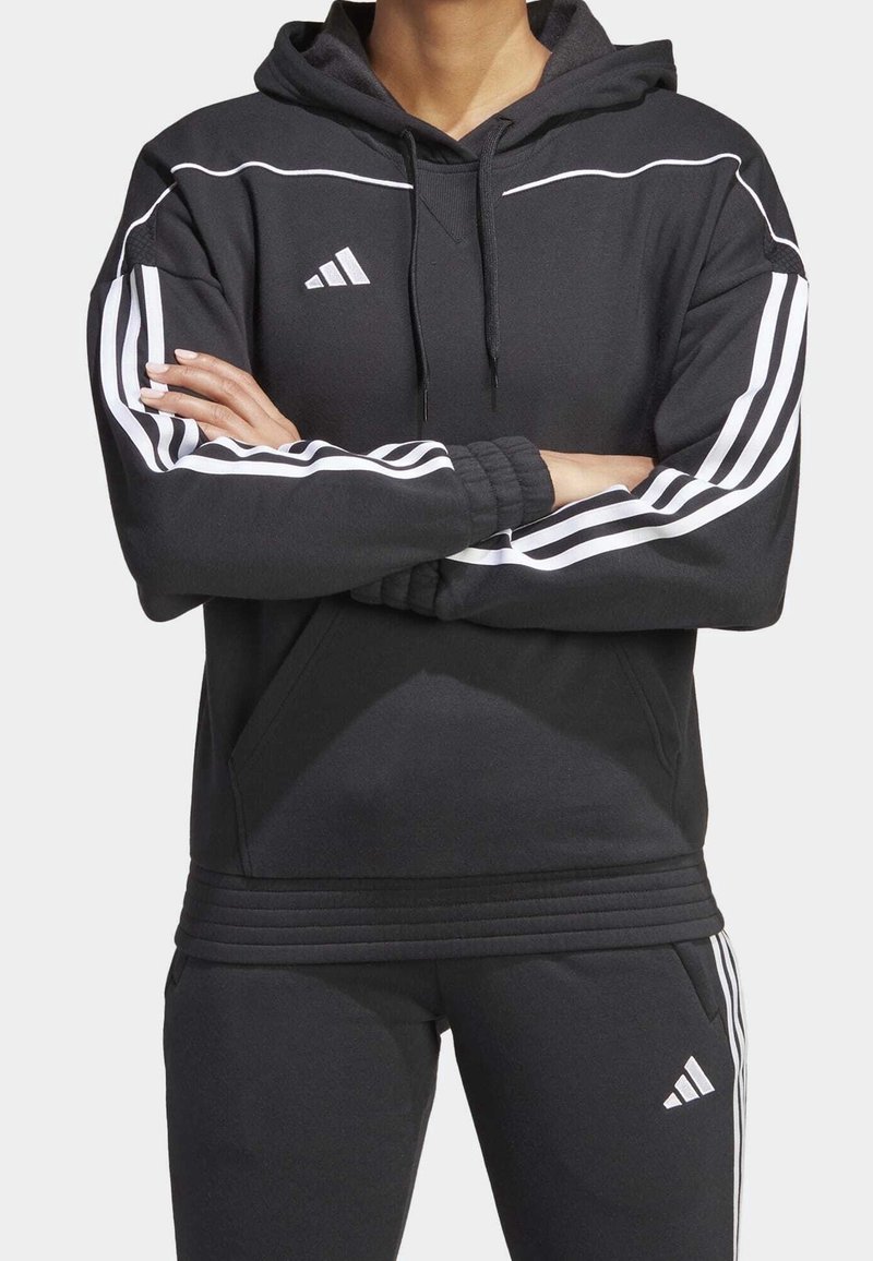 Personne portant un sweat à capuche et un pantalon noirs Adidas avec des rayures blanches, bras croisés, sur un fond clair uni.