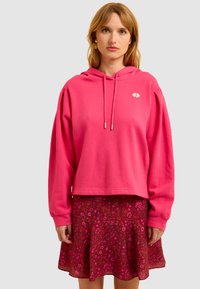 Fuchsia hoodie met een cropped ontwerp en zilveren koordjes, gecombineerd met een bloemig, flared rokje in donkere rode en roze tinten.