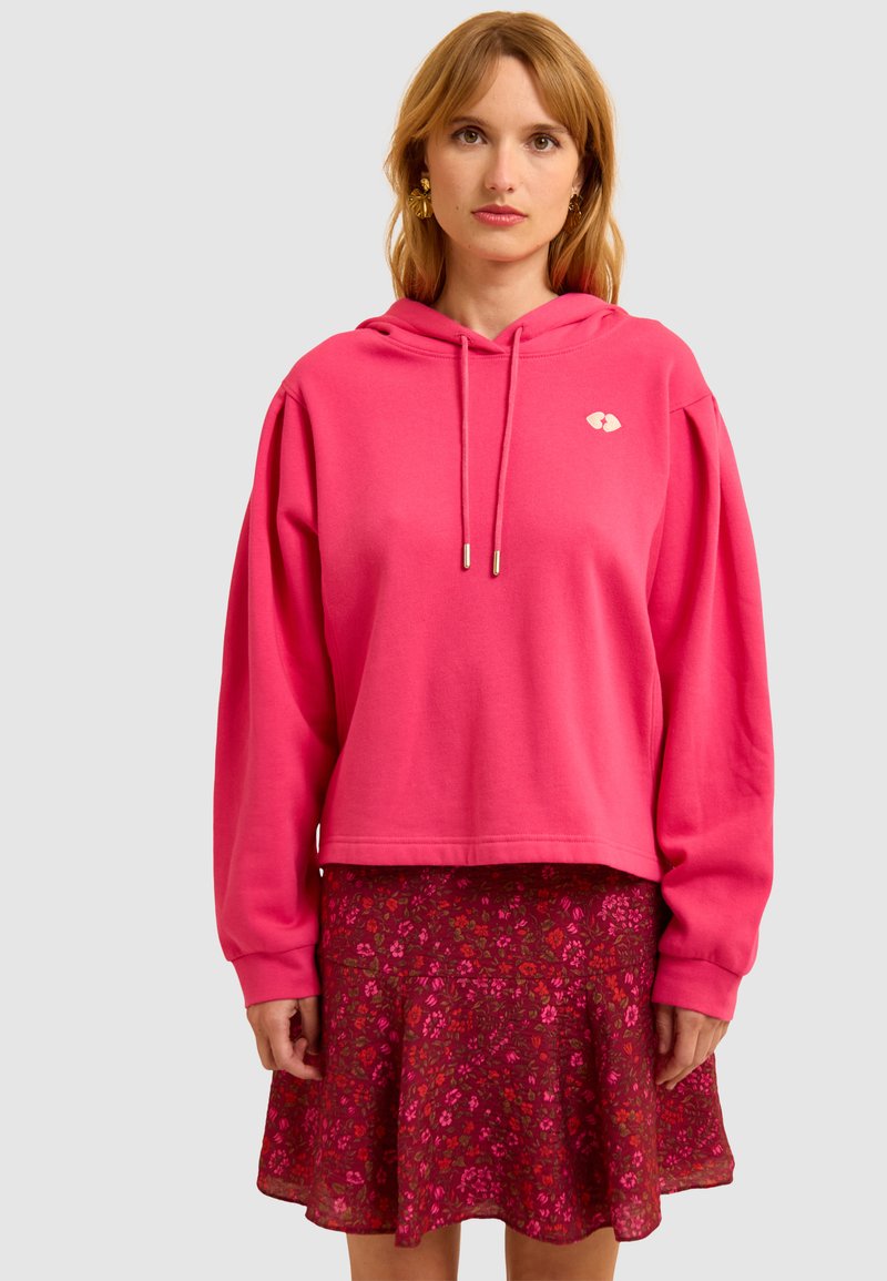 Fuchsia hoodie met een cropped ontwerp en zilveren koordjes, gecombineerd met een bloemig, flared rokje in donkere rode en roze tinten.