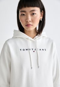 Sudadera blanca de material polar, con un logo de "TOMMYJEANS" en azul marino y un acento rojo. Incluye cordones y un bolsillo tipo canguro.