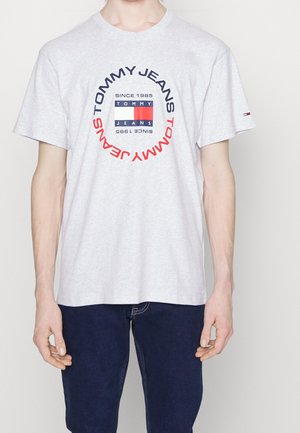 Personne portant un T-shirt gris clair Tommy Jeans avec un logo circulaire et un jean bleu foncé, debout sur un fond clair.