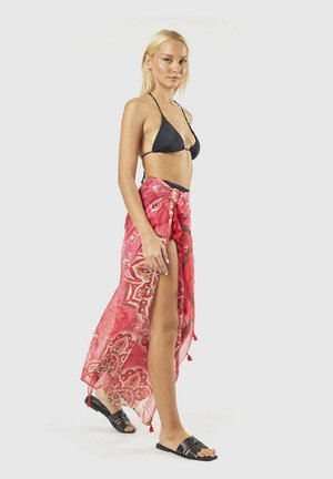 Donna bionda con top bikini nero, sarong rosa con motivi legato in vita e sandali neri, in piedi davanti a uno sfondo semplice.