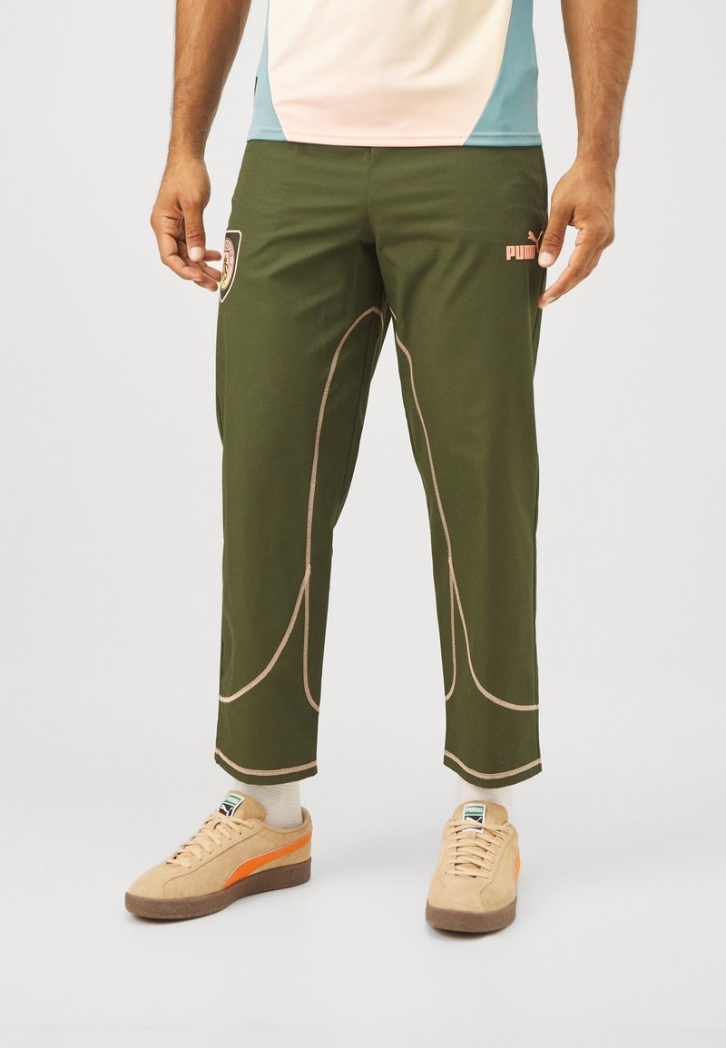 Homme portant un pantalon de sport Puma vert olive avec des coutures roses et un logo écusson, des baskets Puma beiges avec une bande orange, et des chaussettes blanches.