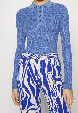 Femme portant un polo bleu côtelé à manches longues et un pantalon blanc taille haute avec de larges rayures abstraites bleues.