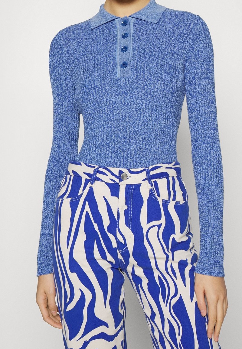 Femme portant un polo bleu côtelé à manches longues et un pantalon blanc taille haute avec de larges rayures abstraites bleues.