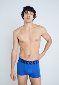 Kék boxer alsónadrág fekete "DIESEL" derékkal, testhez simuló fazonnal és sima textúrával, egy mezítelen tornyú modellen viselve.