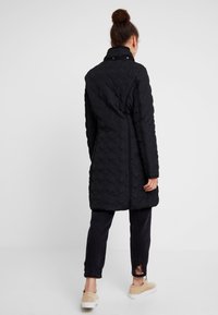 Černý prošívaný kabát s geometrickým vzorem, vysokým límcem a cvoky, kombinovaný s opotřebovanými černými džíny a světlými hnědými botami.