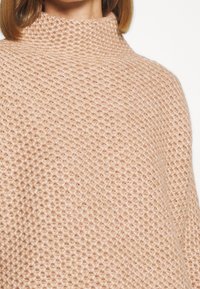 Pull tricoté rose clair texturé avec un col haut. Le tissu présente un motif tissé, offrant chaleur et apparence douce.