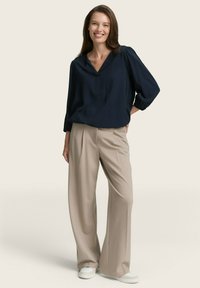 Marineblaue Bluse mit langen Ärmeln, V-Ausschnitt und gerafften Ärmeln, kombiniert mit beigen, weit geschnittenen Hosen und weißen Sneakern.