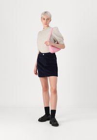 Ανοιχτό γκρι πλεκτό πουλόβερ, σκούρα μπλε κροσέ φούστα, ροζ τσάντα ώμου, μαύρα παπούτσια και κάλτσες. Μοντέλο που στέκεται μπροστά σε λευκό φόντο.