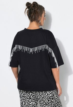 Kvinde med dobbelt knold-frisure bærer sort oversized top med hvide perlekanter på ryggen og sort-hvide mønstrede bukser.
