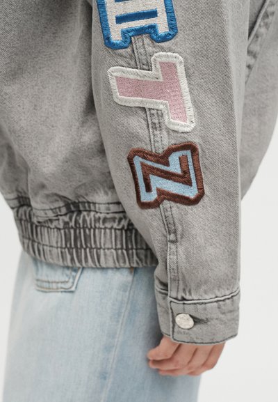 Μανίκι γκρίζου τζιν μπουφάν με πολύχρωμα κεντητά patches και ασημένια μανσέτα με κουμπί, σε συνδυασμό με ανοιχτόχρωμο τζιν.