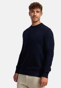 Mørkeblå genser med teksturert herringbone-mønster, rund hals og ribbestrikkede ermer og kant. Laget av et mykt strikket stoff.