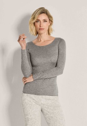 Frau mit kurzen blonden Haaren, die ein graues Langarmshirt und helle Hosen mit Paisley-Muster vor einem schlichten hellen Hintergrund trägt.