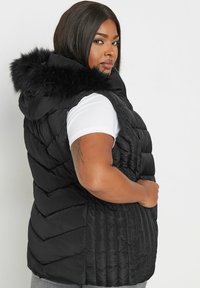 Musta puffaliivi hupulla, jossa on tekoturkistarha, viistokuosiset tikkaat ja vetoketjusuljenta. Puettuna valkoisen paidan päällä.