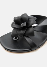 Μαύρο δερμάτινο σανδάλι με φαρδιές λουρίδες και μια μεγάλη μαύρη διακοσμητική ανθοδέσμη στο σημείο του μεγάλου δακτύλου, με ένα μικρό ασημένιο διακοσμητικό στοιχείο.