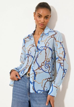 Vrouw die een lichtblauwe blouse met een ketting- en parelprint draagt, gecombineerd met blauwe jeans en gouden sieraden, staand tegen een witte achtergrond.