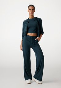 Dunkelblaues Sportset bestehend aus einem Crop-Top und locker sitzenden Hosen. Das Material ist weich und hat eine subtile Textur. Enthält eine Jacke mit Reißverschluss und Taschen.