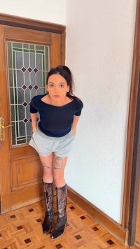 Jeune femme aux cheveux foncés attachés, portant un haut noir, un short clair, des bottes hautes marron brodées, debout à l'intérieur près d'une porte en bois.
