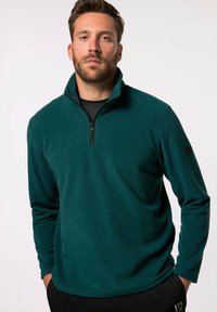 Teal Fleece-Pullover mit viertelzipper Kragen, langen Ärmeln, weicher Textur und einem Logo am linken Ärmel, getragen über einem schwarzen T-Shirt.