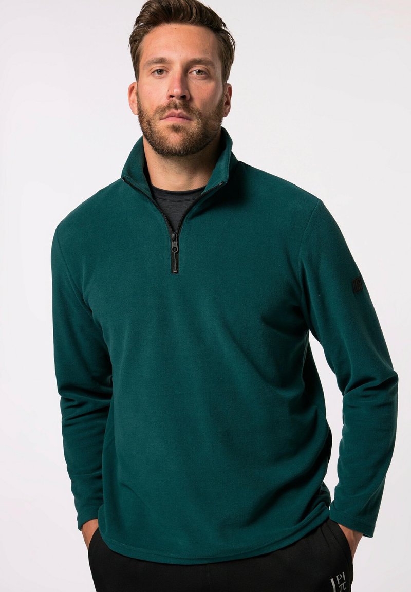 Teal Fleece-Pullover mit viertelzipper Kragen, langen Ärmeln, weicher Textur und einem Logo am linken Ärmel, getragen über einem schwarzen T-Shirt.