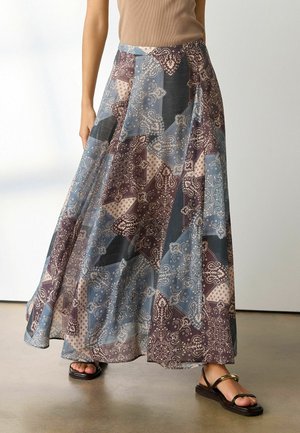 Woman wearing a flowing maxi skirt with blue, brown, and beige patchwork paisley patterns, paired with black flat sandals.