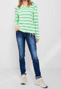 Zielono-biała paskowana bluzka z długim rękawem i dekoltem w serek, zestawiona z niebieskimi dżinsami i jasnymi trampkami. Żyłka przy dolnej części koszuli.