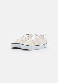 Beige Leinenschuhe mit weißen Schnürsenkeln, schwarzem Sohlenrand und blauen Logodetails. Perforierte Vorderseite für Belüftung, minimalistisches Design.