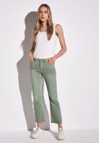 Hellgrüne, gekürzte Jeans mit fransigen Säumen, versehen mit strukturierten Taschen und einem Knopfverschluss. Kombiniert mit einem weißen Tanktop und weißen Sneakers.