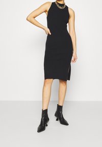 Abito midi nero a coste con scollatura alta, presenta uno spacco laterale. Abbinato a stivali neri a punta. Accento con collana in oro.