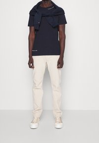 Tmavě modré bavlněné tričko s bílým logem, béžové kalhoty a krémové tenisky. Svetr přehozený přes ramena, hladké textury, neformální střih.