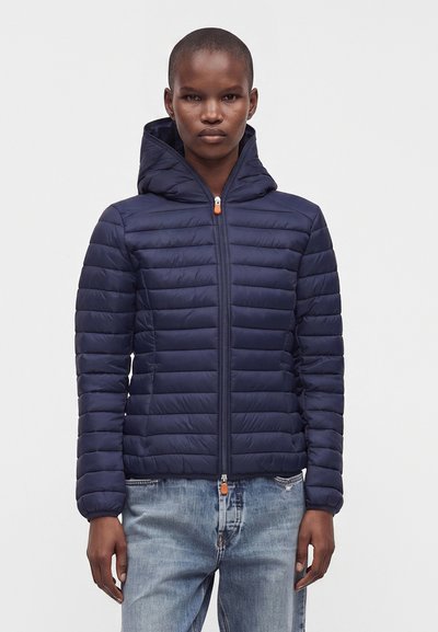Personne portant une doudoune matelassée bleu marine avec capuche et un jean bleu clair, debout devant un fond clair uni.