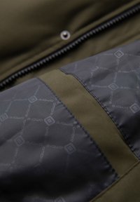 Nahaufnahme einer Jacke, die einen olivgrünen Stoffabschnitt, einen schwarzen Reißverschluss und einen gemusterten dunkelgrauen Stoff mit dem Text "ombre" und Rauten zeigt.